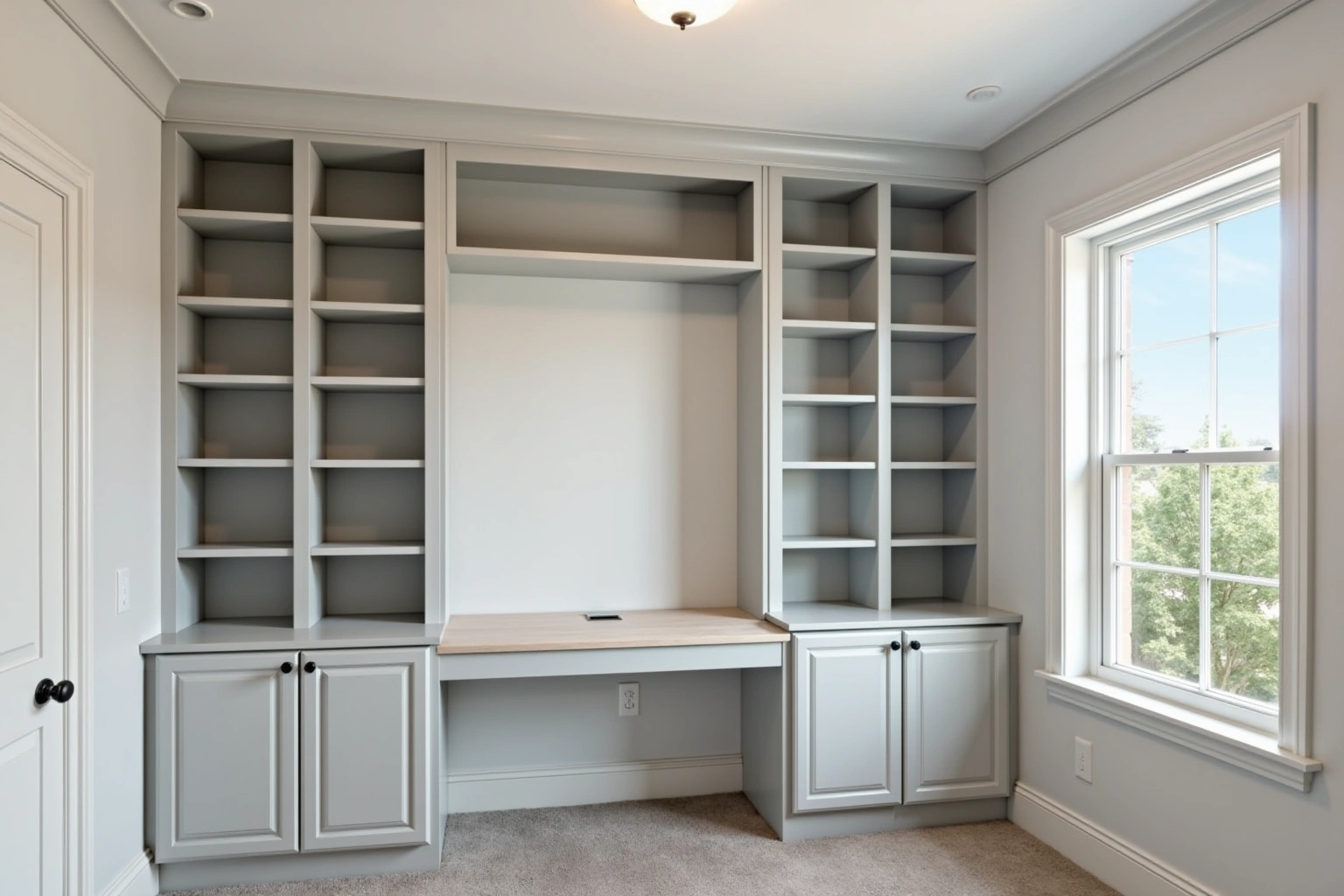 Home office wall of light grey built-in shelves and base cabinets around a wood-top desk beside a window.
